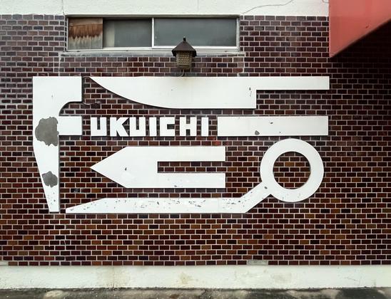 The sign of an old Japanese knife shop. It has a striking design, with the name Fukuichi and some blades of different shapes, on a brick wall.