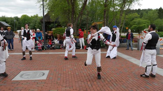 Six morris dancers with white shirts and trousers and black vests, decorated with multicolor ribbons, dancing on a brick paved surface. Four are standing and two are in motion. Behind them an audience including some other morris dancers.