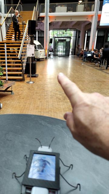 The DigitalBug is on a table overlooking the ZKM lobby. On the right, the KIT Chamber Orchestra is seated, surrounded by the assistance. A big screen above indicates who is speaking. Reinhard's right hand is visible, pointing for the DigitalBug to ZKM director Alistair Hudson speaking from a stair on the left.