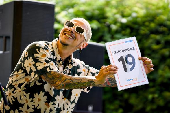 Ein Sänger mit bunt gemustertem Hemd, blondierten Haaren und einer weißgeränderten Sonnenbrille grinst in die Kamera und hält ein Schild mit der Startnummer 19 hoch.
