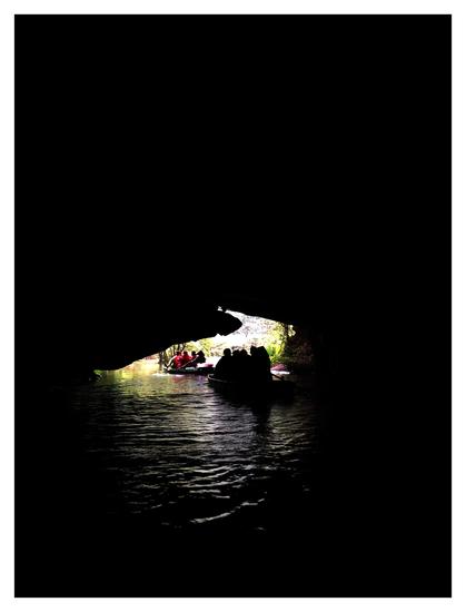 Photo taken on a river in the interior of a partially submerged cave, looking towards a distant jagged-edged opening. A rowboat with passengers sits in silhouette at the opening, while another boat ahead of it is fully in the light. A green jungle can be glimpsed beyond, and reflected sunlight plays on the river’s rippling waves. The rest of the image is plunged in darkness.