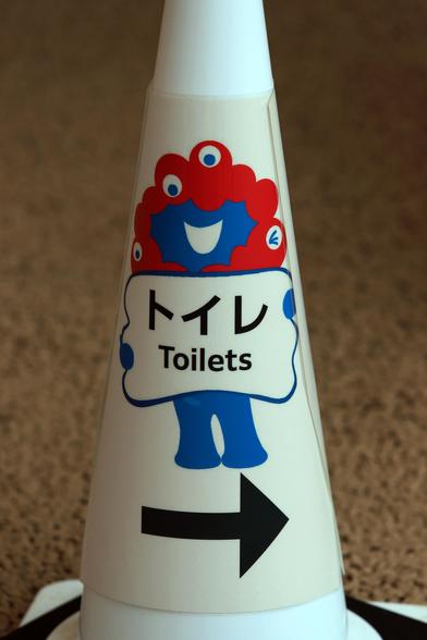 Ein Straßenhütchen mit dem blau-roten und mit Augen bedeckten Expo-Maskottchen. Es hält ein Schild in der Hand und weisst den Weg zu den Toiletten.