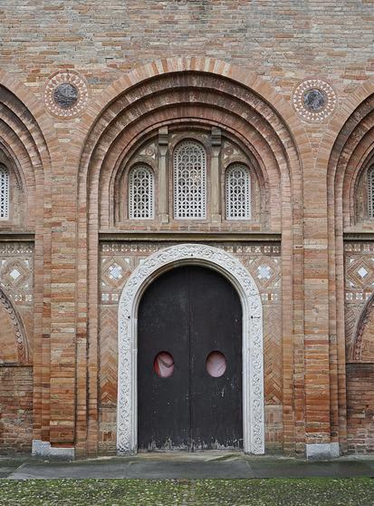 Porte ancienne en bois banale, mais avec autour, dans l’ordre : un encadrement en pierre blanche délicatement ouvragée,m ; une embrasure en brique avec au niveau des écoinçons des motifs géométriques en brique et pierre ; au-dessus, un tympan en brique aéré par trois ouvertures verticales également en pierre ouvragée ; tout autour encore des archivoltes en briques qui encadrent l’ensemble et lui donnent de la profondeur.