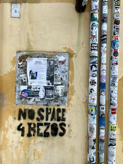 Colour photo of a stained and weather worn ochre yellow wall, with three narrow pipes running down the right hand side of the frame, covered in stickers. On the left is a square of metal, it too covered in stickers and half torn-off posters. Underneath, in emphatic heavy black capital letters is written "no space 4 Bezos"