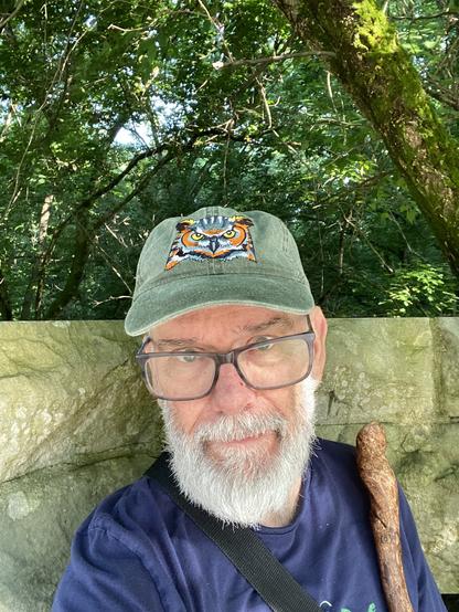 Selfie with owl cap at "Mildred" bridge with stone railing behind me, walking stick on shoulder