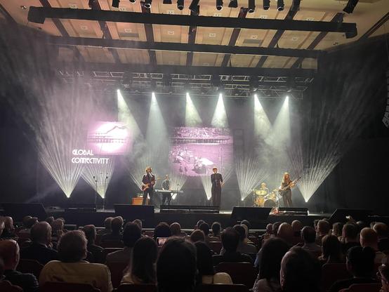 A live music performance on stage with a band featuring a singer and musicians playing keyboard and guitar. The backdrop displays the text "GLOBAL CONNECTIVITY" along with visual effects and lighting. An audience is seated in the foreground, watching the performance.