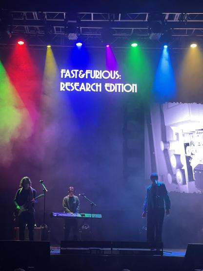 A live performance scene featuring three musicians on stage. One plays an electric guitar, another is on a keyboard, and the third is singing. Colorful lights illuminate the stage with a backdrop displaying the text "FAST & FURIOUS: RESEARCH EDITION”