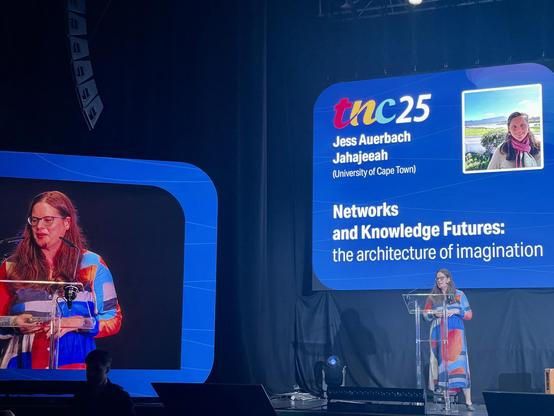 Jess Auerbach Jahajeeah, a woman with long hair and glasses, is wearing a long bright and colourful dress, stands at a podium, speaking at a conference. A large screen behind her displays her name, affiliation with the University of Cape Town, and the title of her presentation: "Networks and Knowledge Futures: the architecture of imagination”