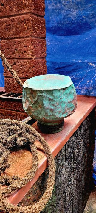 A heavily rented and oxidized-green copper pot placed upside down on the wall of a well. Red walls. A thick drawing rope in the left foreground. Blue tarp in the background.