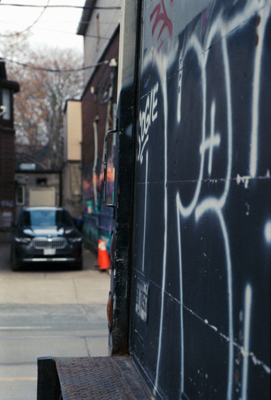 The rear deck of a well-worn delivery truck has been tagged. Not for the first time, as the truck owner has clearly painted over tags in the past. Across the street, the line of the truck deck is continued by a colourful wall of street art but it is out of focus.