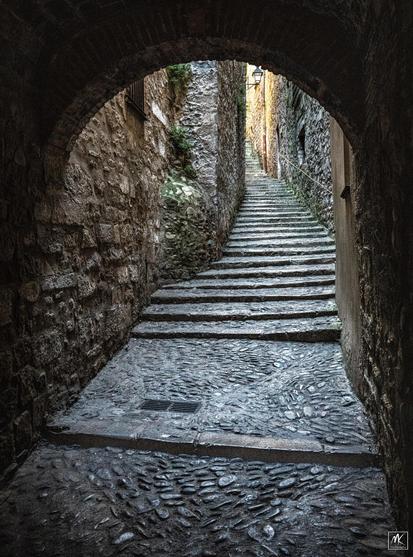 Color photo looking through a dark rounded arch toward a narrow passageway of stone steps winding upward.