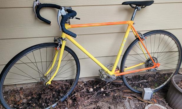 Orange and yellow bike, black wheels, black seat, dark green handlebar tape, against a wood siding house