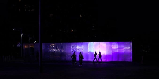 A smooth, sleek wall panaled in metal sheet reflects violet light. Against the light we see the silhouettes of two women walking togehter, plus another woman walking alone and project a perfect shadow on the wall which resembles a ghost.
