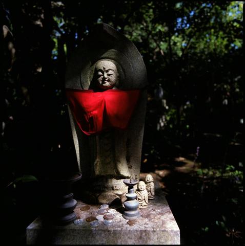 a small japanese buddhist statue in a pool on sunlight. the statue is wearing a red gape, which is very vivid in the sun. offerings are places in front of the status.