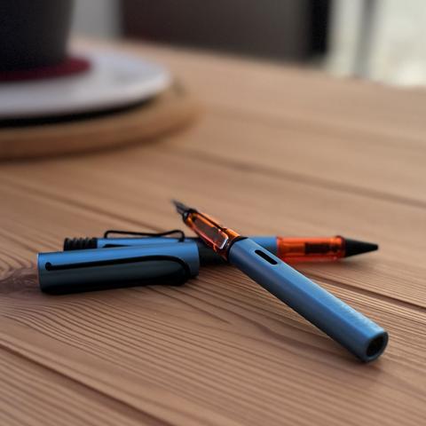 Sobre mesa de madera, cruzadas, están el boli y la pluma, de cuerpo azul grisáceo y detalle en naranja.