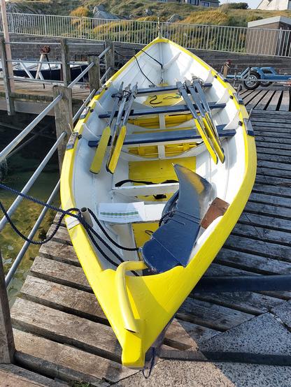 A coastal rowing yoal in yellow, white and blue on land.