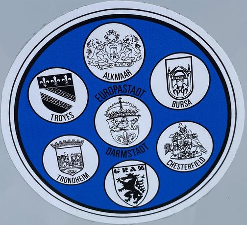 Runder Sticker mit blauem Hintergrund. In der Mitte kreisrund das Wappen der Stadt Darmstadt in weiß und den Worten „Europastadt“ oberhalb und „Darmstadt“ unterhalb. Drumherum gruppiert, ebenfalls kreisrund die Wappen der Partnerstädte Trondheim, Graz, Chesterfield, Bursa, Alkmaar und Troyes.