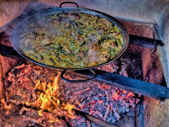 La imatge mostra una paella cuinant a foc obert. La paella, un plat d'arròs tradicional valencià es troba en una paella gran i rodona, i l'arròs es cuina amb pollastre i mongetes verdes. El foc és de fusta, i les flames són visibles sota la paella. El vapor surt de la paella, indicant que fa calor i acabat de fer.