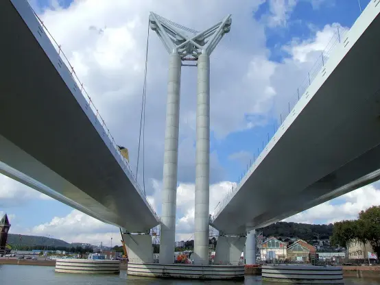 The image shows a modern bridge with a distinctive design, featuring two tall concrete pillars supporting the bridge deck. The bridge has a sleek and contemporary appearance, with cables likely providing additional support. The background includes cloudy skies and possibly a body of water underneath the bridge, along with some buildings and structures in the vicinity. Overall, it captures an impressive engineering feat.

Image Credits: Wikimedia / MOSSOT / CC BY 3.0