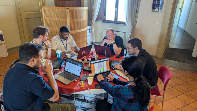 Group of 5 students sitting together with tutor Sebastian Rudolph from TU Dresden around a table, stuffed with computing infrastructure, thinking....