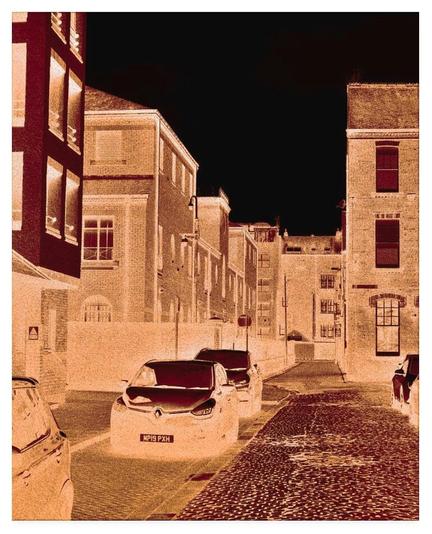 Colour modified (mainly a russet colour with a black sky) portrait photograph showing the approach to a narrow lane with cars parked on either side of a cobbled road.