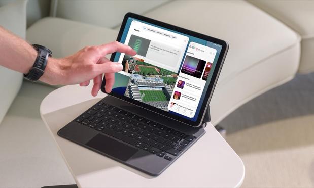 Eine Hand bedient ein iPad samt angebrachtem Magic Keyboard von Apple mit der neuen Multitasking-Funktion. Das iPad steht auf einem kleinen weißen Tisch, im Hintergrund eine weiße Sofalandschaft.