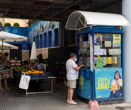 A street scene in Palafrugell, Spain, showing a man in shorts and a white t-shirt purchasing lottery tickets from a blue ONCE lottery kiosk adorned with posters and scratch cards, next to an open-air market stall selling fresh produce under white umbrellas. The kiosk features the text "BEN JUGAT" and is topped with a white canopy, while the background includes a blue mural and market shoppers.