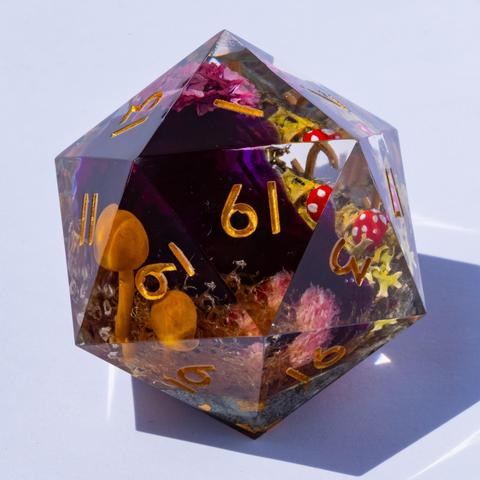 A oversized D20 with a diorama featuring moss, small mushrooms and a purple skull on white background. Inked in gold.