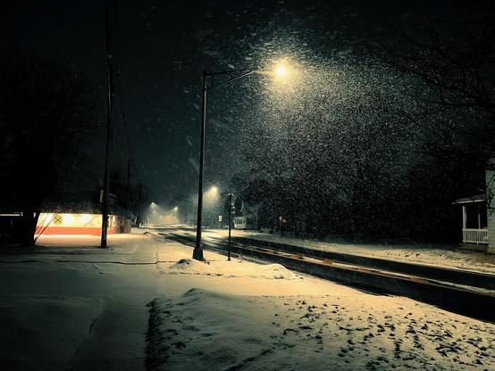 A snowy street at night is dimly lit by streetlights, with snow falling heavily and blanketing the ground, trees, and houses. A warmly lit building glows on the left side, adding contrast to the cold, dark scene.