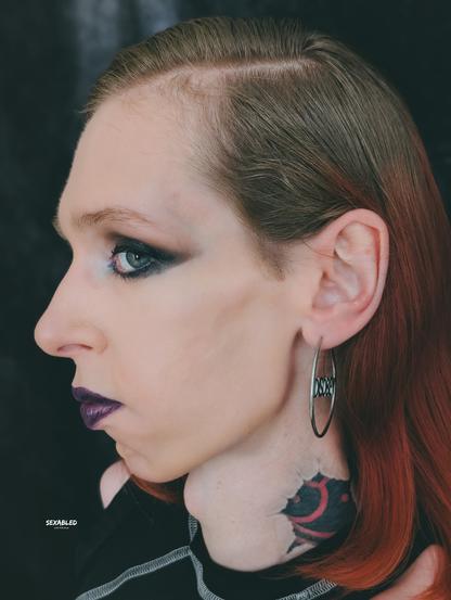 Ein Porträt von mir Profil mit langem, rotbraunem Haar, das nach hinten gekämmt ist. Ich trage dunkles Augen-Make-up und lila Lippenstift. Am Ohr hängt ein großer, silberner Creolen-Ohrring, in dem "Disobey" steht. Am Hals ist mein BDSM-Tattoo sichtbar. Der Hintergrund ist dunkel.