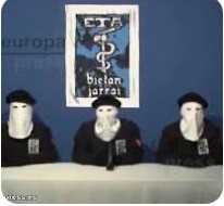 Three individuals wearing black clothing and white masks are seated at a table in front of a blue background. A banner with the letters "ETA" is displayed above them.