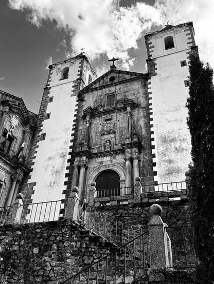 Fotografía en blanco y negro de una imponente iglesia o monasterio. La imagen se centra en la fachada principal, mostrando una escalinata de piedra que conduce a una entrada principal con un arco de medio punto.  A ambos lados de la entrada principal se alzan dos torres altas y estrechas, con una textura que sugiere antigüedad. La fachada principal está elaborada, con detalles arquitectónicos como columnas, nichos y un escudo. En la parte superior, una cruz se alza sobre el edificio. Al fondo se puede apreciar una parte lateral del complejo, y a la derecha, un ciprés. La escena está tomada desde una perspectiva baja, acentuando la altura y la grandeza de la construcción.