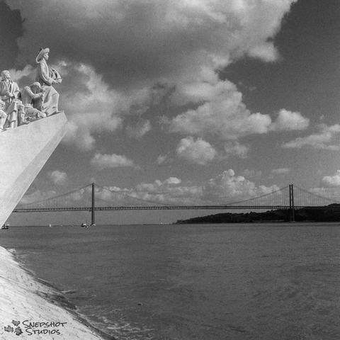 Square black and white photo. A large suspension bridge in the background and a stone monument with statues partially cut off at the left side
