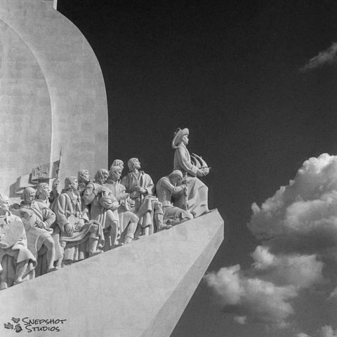 Square black and white photo. A stone monument with statues of humans on the left. The right side shows the sky with a few white clouds