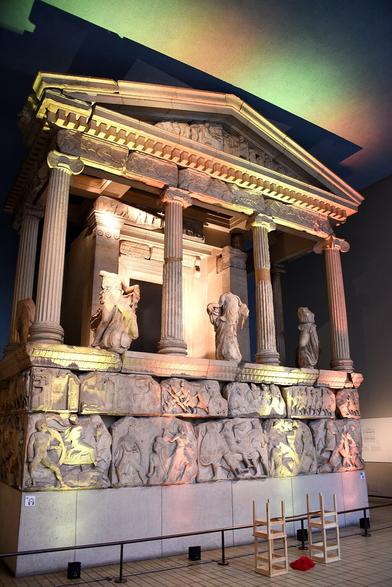 Photograph of the Nereid Monument that is in the British Museum. The structure has a traditional Ancient Greek style with a triangular roof and columns. Three carved figures are between the front four columns Below them is a fresco with figures carved across it.