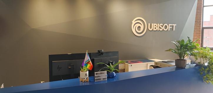 A picture of Ubisoft's front desk. A typical front desk with the Ubisoft logo on the wall. It has a pride flag on the desk.