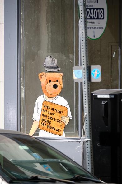Wheatpaste graffiti of a man with a teddy bear for a head, holding a sign for the Step Outside Art Show.