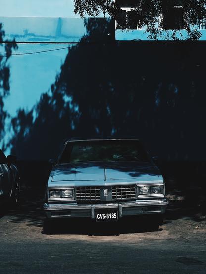 A vintage car with a front view is parked in the shade. The car is light blue and has a black license plate reading "CVS-8185." Shadows from nearby trees are cast on the vehicle and the bright blue wall behind it, creating a pattern of light and dark on the surfaces. The setting appears to be outdoors on a sunny day.