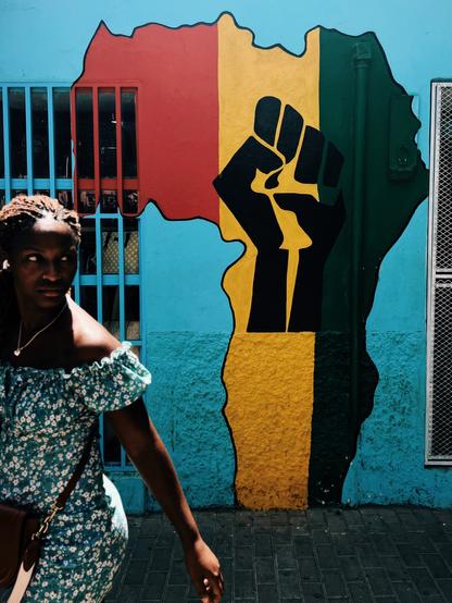 A person in a blue floral dress walks past a colorful mural. The mural features a raised fist symbol within the outline of the African continent, painted in red, yellow, and green stripes. The background is a vibrant blue with metal bars visible on the left side.