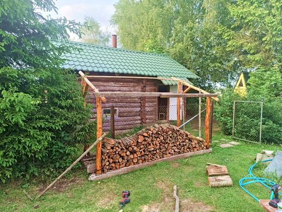 A small firewood store is taking shape. It straddles a wire fence. There's four concrete beams recycled from decomssioned power pylons as base on which the firewood is stacked off the ground. Then there's four corner posts made from debarked logs and two beams connecting them across the fence, with a diagonal on either side for stability. The two sides are temporarily joined by some scrap wood pieces to keep them aligned. A temporary brace is supported by a stick hammered in the ground.