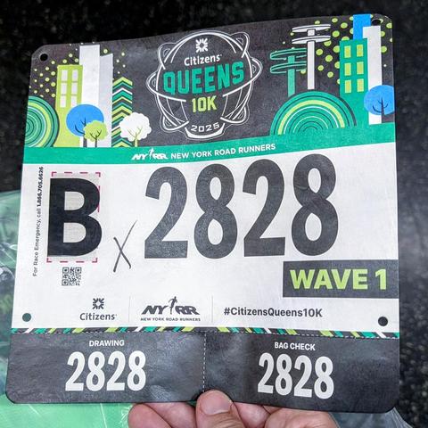 A hand holding a bib for a road running race. It has a logo saying "Citizens Queens 10k", the number "B 2828", and a label saying "wave 1" in it. Logos of the sponsors and organizers (Citizens, NY Road Runners) are in the bottom.