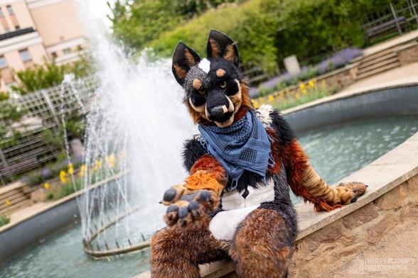 Blue Heeler fursuiter sitting on the edge of a fountain, offering a paw to the viewer, inviting them to sit down next to him.