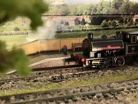 A view across a small model railway scene at a platform at the back with a small black steam engine in front of it. On the platform is Paddington in his blue coat, red hat and holding a suitcase
