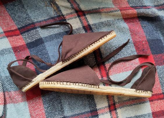 A pair of brown linen espardilles on a blue and red checkered blanket. The shoes have straps attached to the heel to keep them on feet.