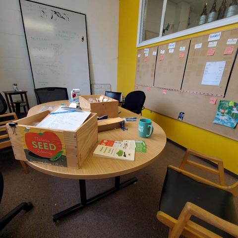 A brightly lit meeting room features a round wooden table surrounded by chairs. On the table are a wooden box labeled "Victoria Seed Library," a stack of "Welland Community Orchard" flyers, and a turquoise mug that says "But first, Coffee". In the background, a whiteboard covered with handwritten notes and drawings stands against a white brick wall. A corkboard divided into months with notes is on a yellow wall next to a window. The room appears to be in an office or community space.
