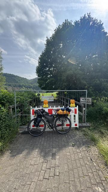Ein Fahrrad ist vor einer geschlossenen Barriere auf einem von Grün umgebenen Weg geparkt. Die Schranke hat Warnleuchten und es gibt ein Schild, das auf den eingeschränkten Zugang hinweist. Im Hintergrund sind Hügel und ein bewölkter Himmel zu sehen.
