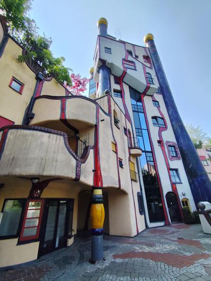 Das Foto zeigt ein außergewöhnliches, farbenfrohes Gebäude mit organischen Formen und verspielter Architektur. Die Fassade ist unregelmäßig geschwungen, ohne gerade Linien oder rechte Winkel. Das Gebäude besteht aus mehreren Etagen und besticht durch eine Kombination aus kräftigen Farben wie Gelb, Rot, Blau und Schwarz.

Besonders auffällig sind die beiden hohen, blau gefliesten Säulen mit goldenen Kuppeln an der Spitze, die wie Türme wirken und dem Bau ein märchenhaftes Aussehen verleihen. Die Fenster sind verschieden groß und in unregelmäßiger Anordnung verteilt, was dem Gebäude einen verspielten, fast surrealen Charakter verleiht. Viele Details wie Mosaike, gewellte Fassadenlinien und unterschiedlich geformte Balkone tragen zur einzigartigen Gestaltung bei.