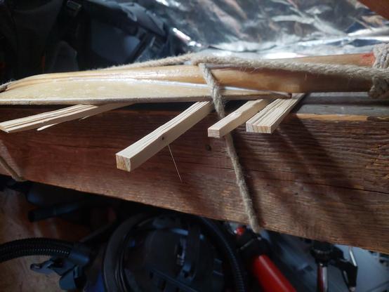 Ein Holz-Paddel ist auf einen Balken gebunden. Keile stützen die gebogene Form, sodass der Leim zwischen Brett und Griff gut aushärten kann.