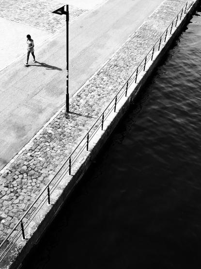 a hot sunny day. Frankfurt. Bank of the Main. Photographed diagonally downwards from a bridge. The photo is strictly diagonally divided into two parts. on the right the dark, almost black water surface. on the left the bright asphalted and paved riverbank promenade. a single passer-by on the sidewalk casts a hard shadow on the asphalt. i omit the ugly lantern that has no function in the picture in the description. it was just standing there :)