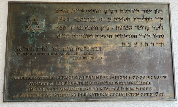 Das Bild zeigt eine Gedenktafel an einer Wand. Die Tafel ist aus Metall und hat eine grünlich-braune Patina. Sie enthält Inschriften in hebräischer und deutscher Sprache sowie ein Davidstern-Symbol.  Die hebräische Inschrift ist in mehreren Zeilen oben auf der Tafel zu sehen. Darunter folgt eine deutsche Inschrift, die lautet:  "Gedenke, ewiger, was an uns geschah... (Klagelied 5,1)  An dieser Stelle befand sich die in den Jahren 1827-28 erbaute Synagoge der jüdischen Gemeinde Wattenscheid. In der Pogromnacht des 9./10. November 1938 wurde sie durch Brandstiftung der Nationalsozialisten zerstört."  Die Tafel erinnert an die Zerstörung der Synagoge während der Novemberpogrome 1938 und dient als Mahnmal für die Opfer der nationalsozialistischen Gewaltherrschaft.